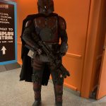 A full-armor Mandalorian cosplayer in beskar helmet and cape holding a blaster rifle at a convention.