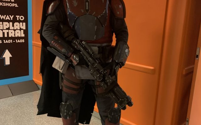 A full-armor Mandalorian cosplayer in beskar helmet and cape holding a blaster rifle at a convention.