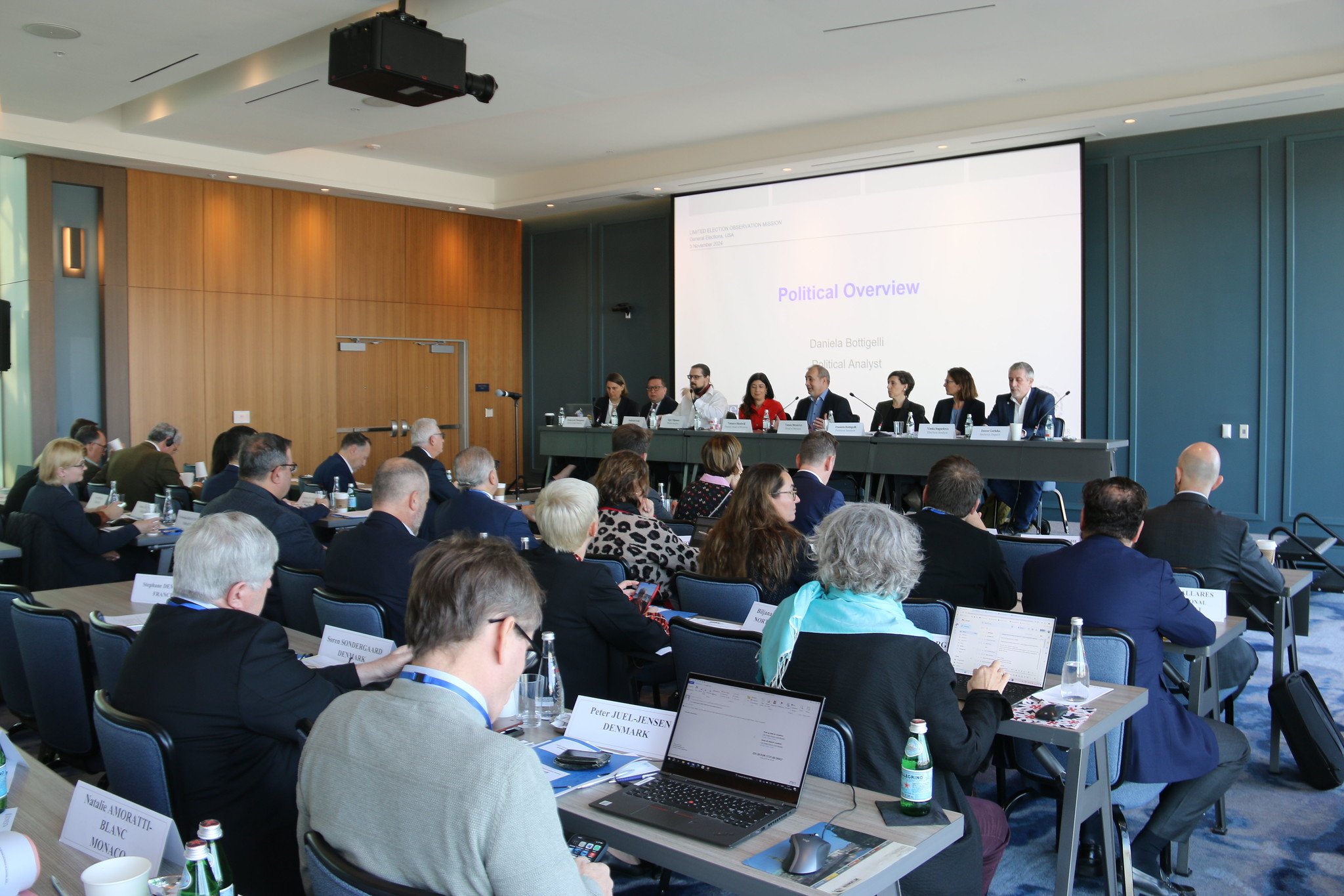Rows of observers at desks with laptops and country name cards face a panel of eight ODIHR staff under a screen reading Limited Election Observation Mission, General Elections, USA, 5 November 2024