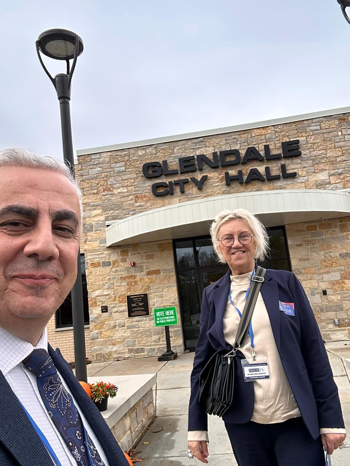 Two OSCE observers wearing blue lanyards and badges stand outside the stone front of Glendale City Hall, with a green Vote Here sign by the door