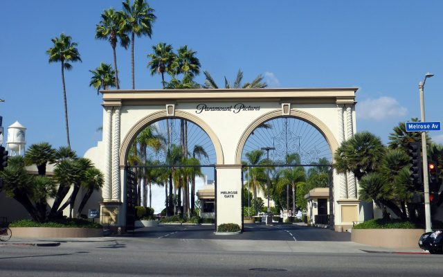 The cream double-arched Paramount Pictures Melrose Gate on Melrose Avenue in Hollywood under a clear blue sky, with palm trees and a studio water tower behind it