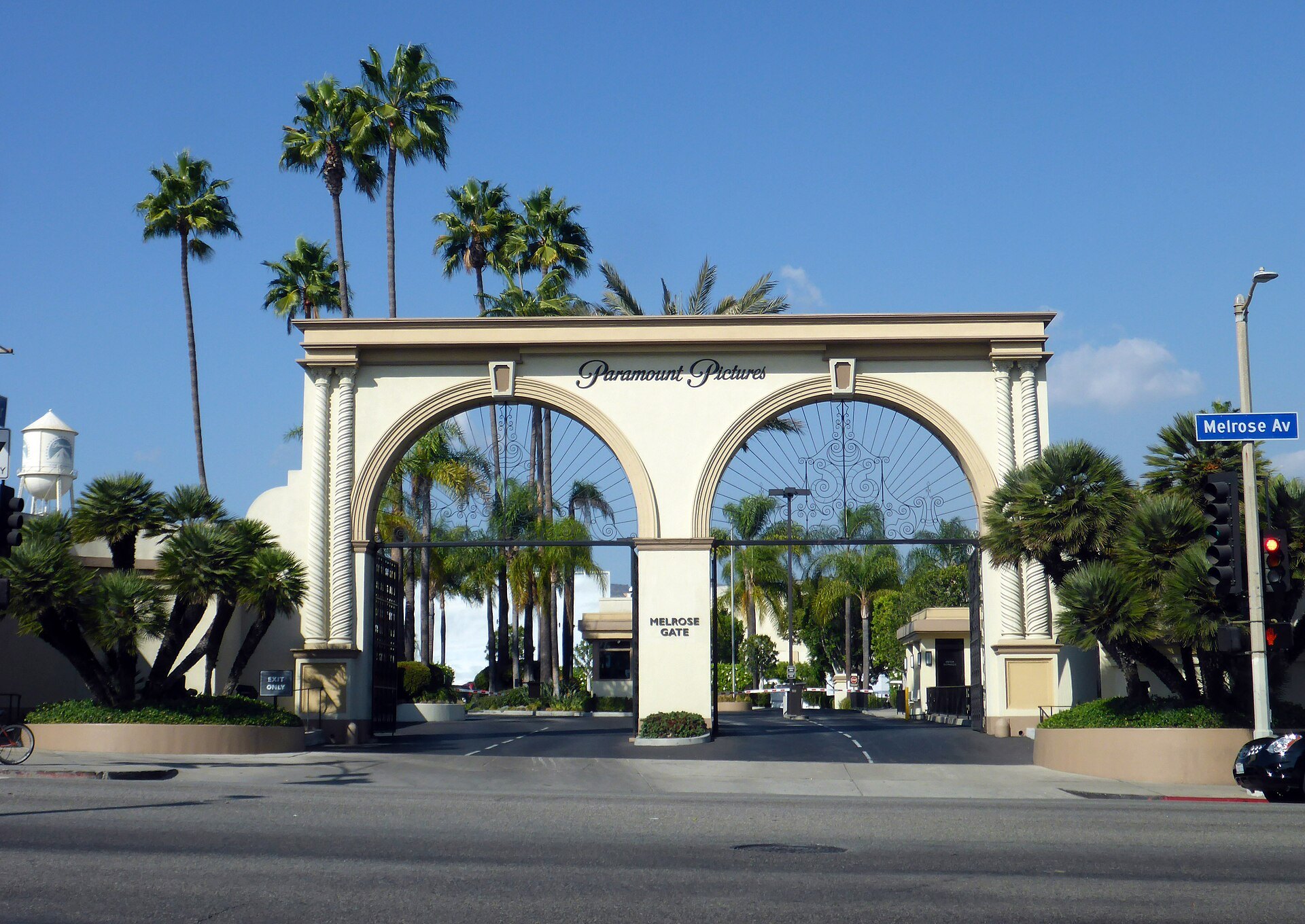 The cream double-arched Paramount Pictures Melrose Gate on Melrose Avenue in Hollywood under a clear blue sky, with palm trees and a studio water tower behind it