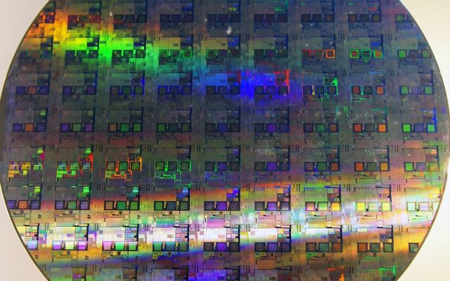 A circular silicon wafer patterned with a grid of identical microchips, catching rainbow light.