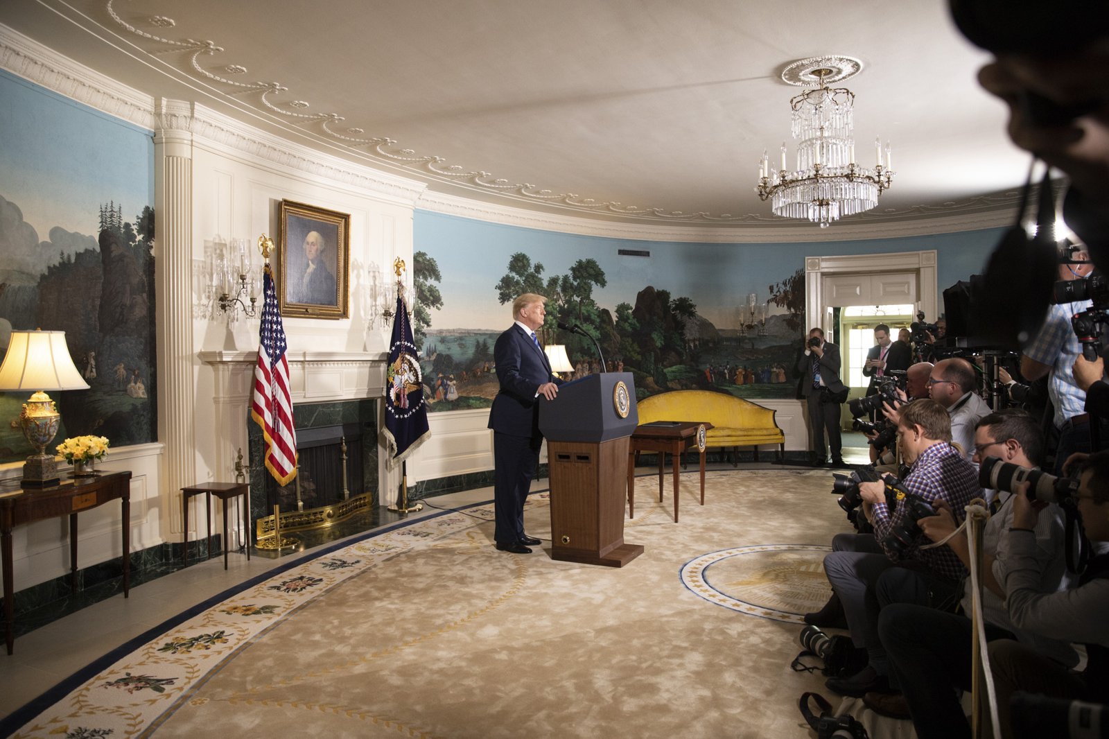President Donald Trump speaks at a lectern in the White House Diplomatic Reception Room as photographers work nearby