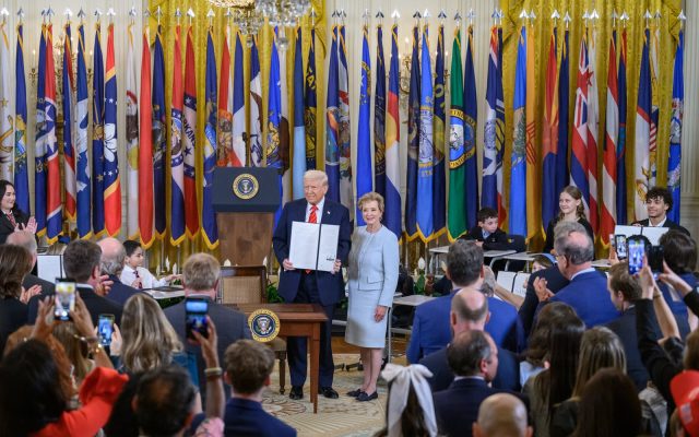 President Donald Trump holds up a signed executive order beside Education Secretary Linda McMahon in the East Room, with schoolchildren at desks and state flags behind them