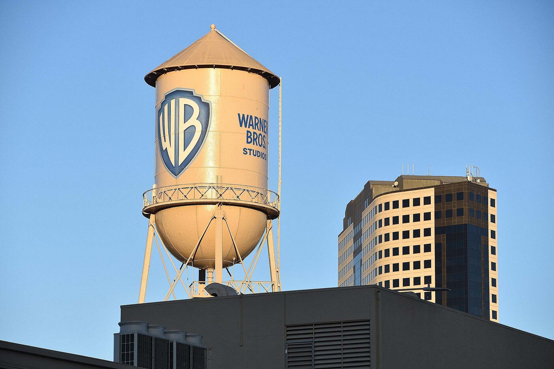 The Warner Bros. Studios water tower with its blue WB shield, lit by warm light against a blue sky, beside an office tower in Burbank