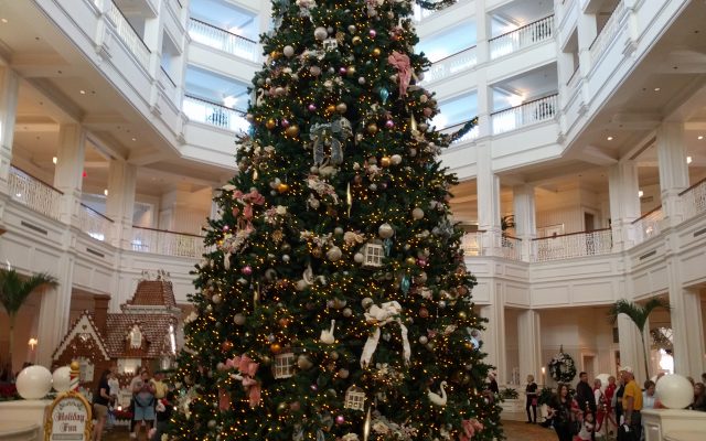 A tall decorated Christmas tree in the white atrium lobby of a resort, with a gingerbread house at its base