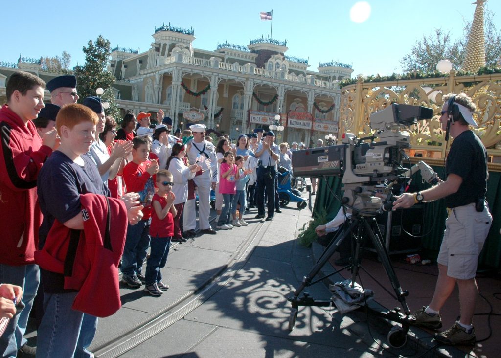 A television camera on a tripod faces a row of guests and sailors in dress uniform clapping along Main Street at Magic Kingdom, with holiday garlands on the buildings
