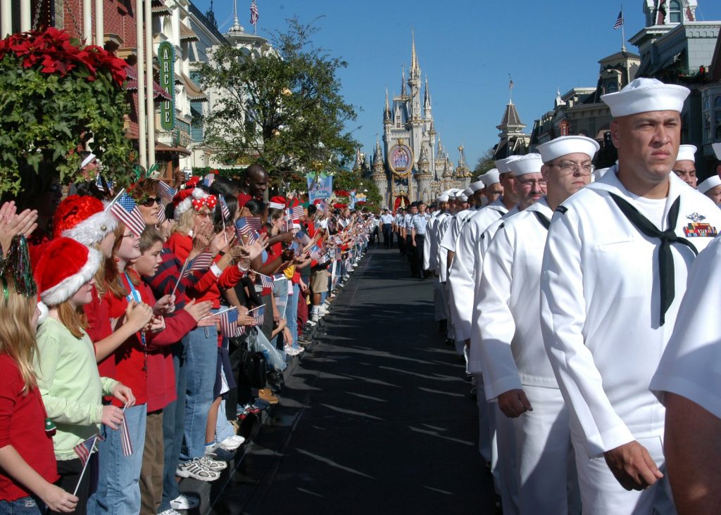 Sailors in white dress uniforms march down Main Street toward Cinderella Castle past guests waving small American flags