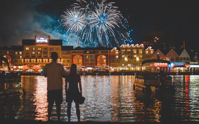Two people silhouetted at the edge of Crescent Lake watching fireworks over the lit-up BoardWalk at night