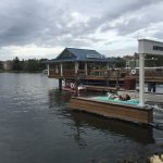 A turquoise vintage Amphicar with a captain and passengers splashes into the lake beside the Amphicar Launch sign at The Boathouse, Disney Springs