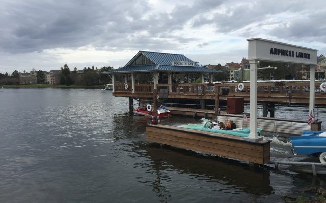 A turquoise vintage Amphicar with a captain and passengers splashes into the lake beside the Amphicar Launch sign at The Boathouse, Disney Springs