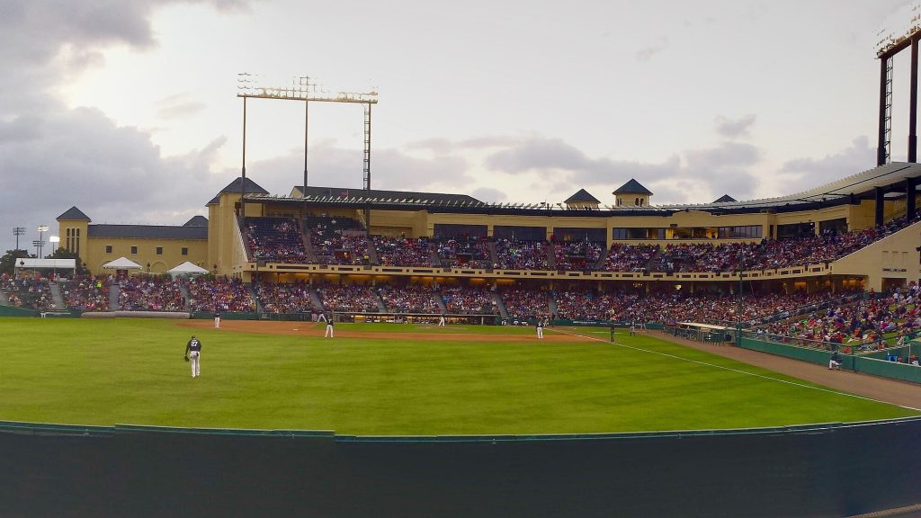 Wide view of a baseball stadium at dusk, with a covered grandstand full of fans behind a green outfield and a tall light tower overhead.