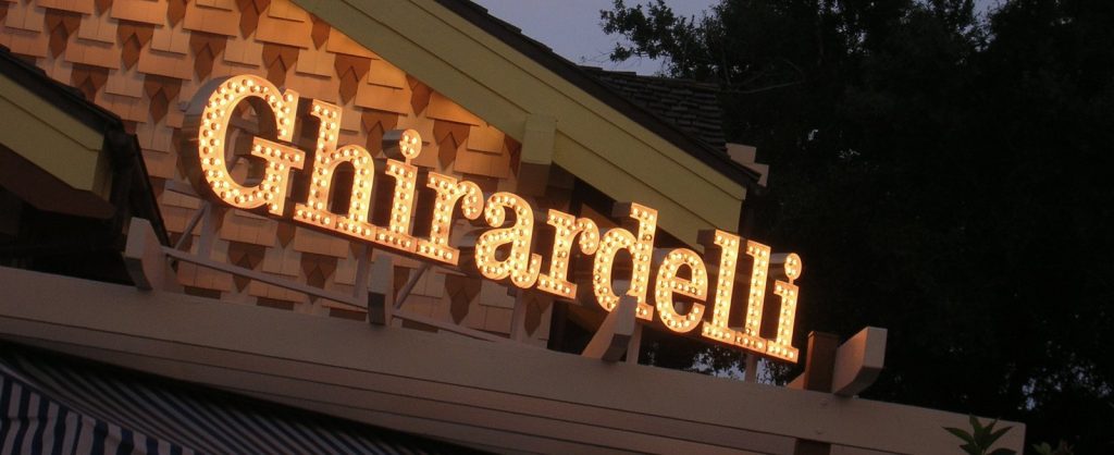The lit marquee-bulb Ghirardelli sign on the roof of the Marketplace shop at dusk