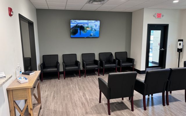 An empty doctor's office waiting room with black armchairs along a gray wall and a television mounted above them