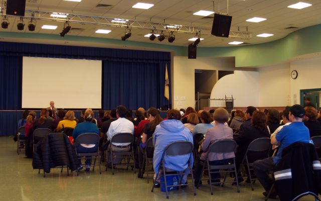 Residents seated on folding chairs in a community hall, seen from behind, listening to a presentation on Affordable Care Act coverage
