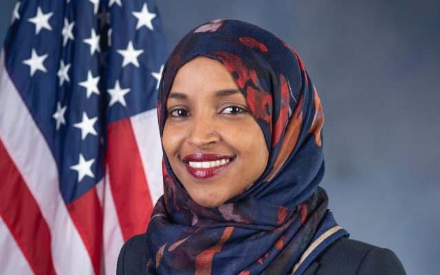Rep. Ilhan Omar smiling in a patterned navy and red head scarf and dark jacket in front of an American flag