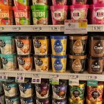 A supermarket freezer shelf stacked with colorful ice cream cartons, each shelf tag showing a price and a price per ounce