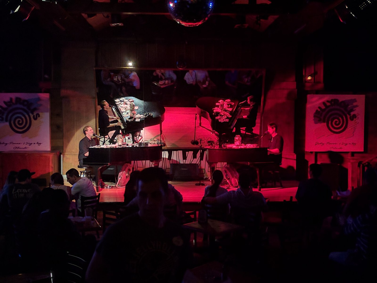 Two pianists on the red-lit stage at Jellyrolls with a crowd at tables in the foreground