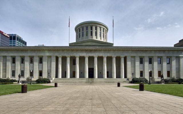 The limestone Ohio Statehouse in Columbus beneath a blue sky, with its columned west front facing the lawn.