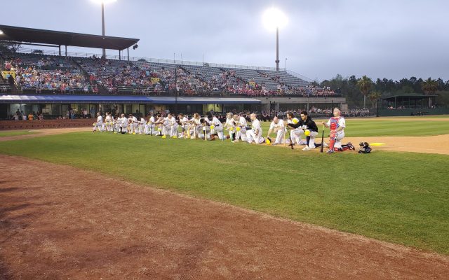 Wide view of a ballpark at dusk with a row of players in white uniforms kneeling along the infield grass in front of a grandstand of fans.