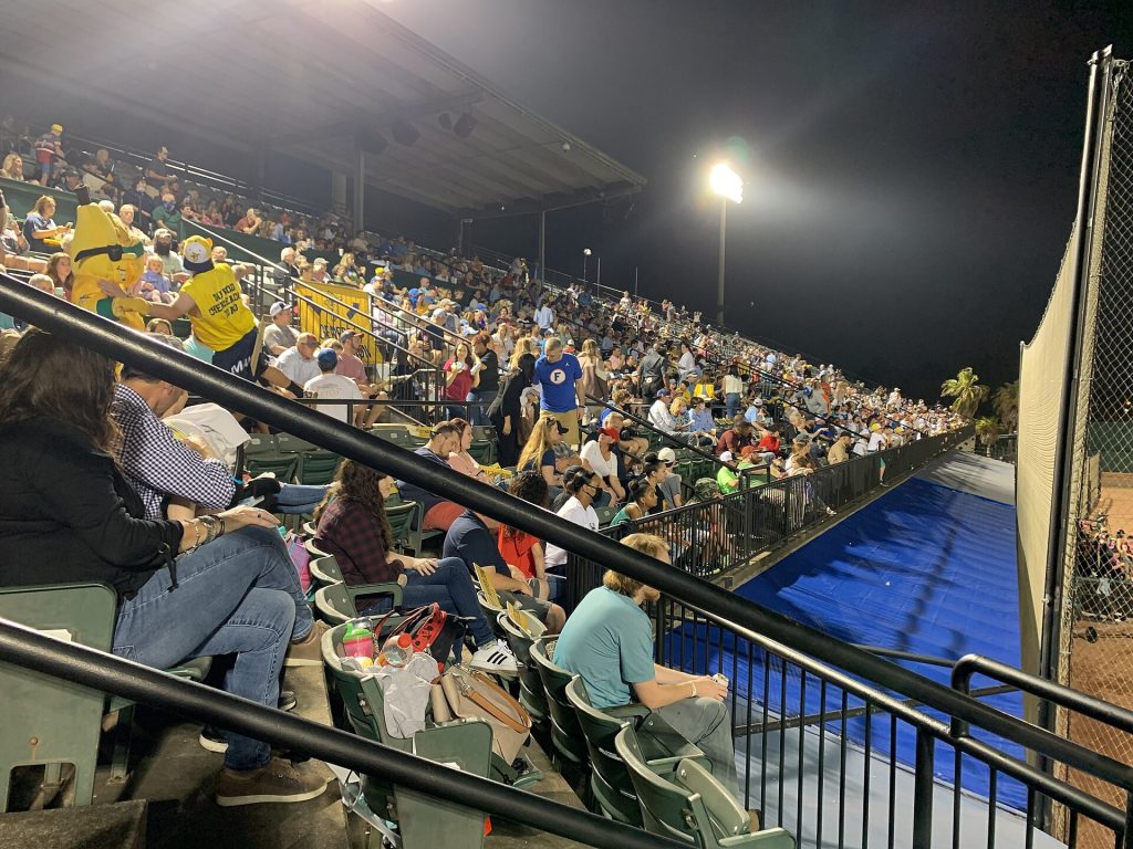 Fans in a covered grandstand at night, with a person in a yellow banana costume among them and a blue tarp at right.