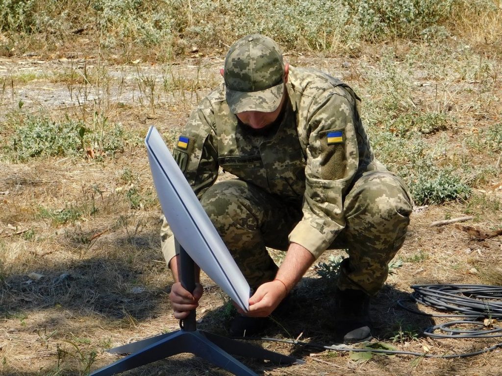 A Ukrainian soldier sets up a white Starlink satellite dish on its stand in a dry field